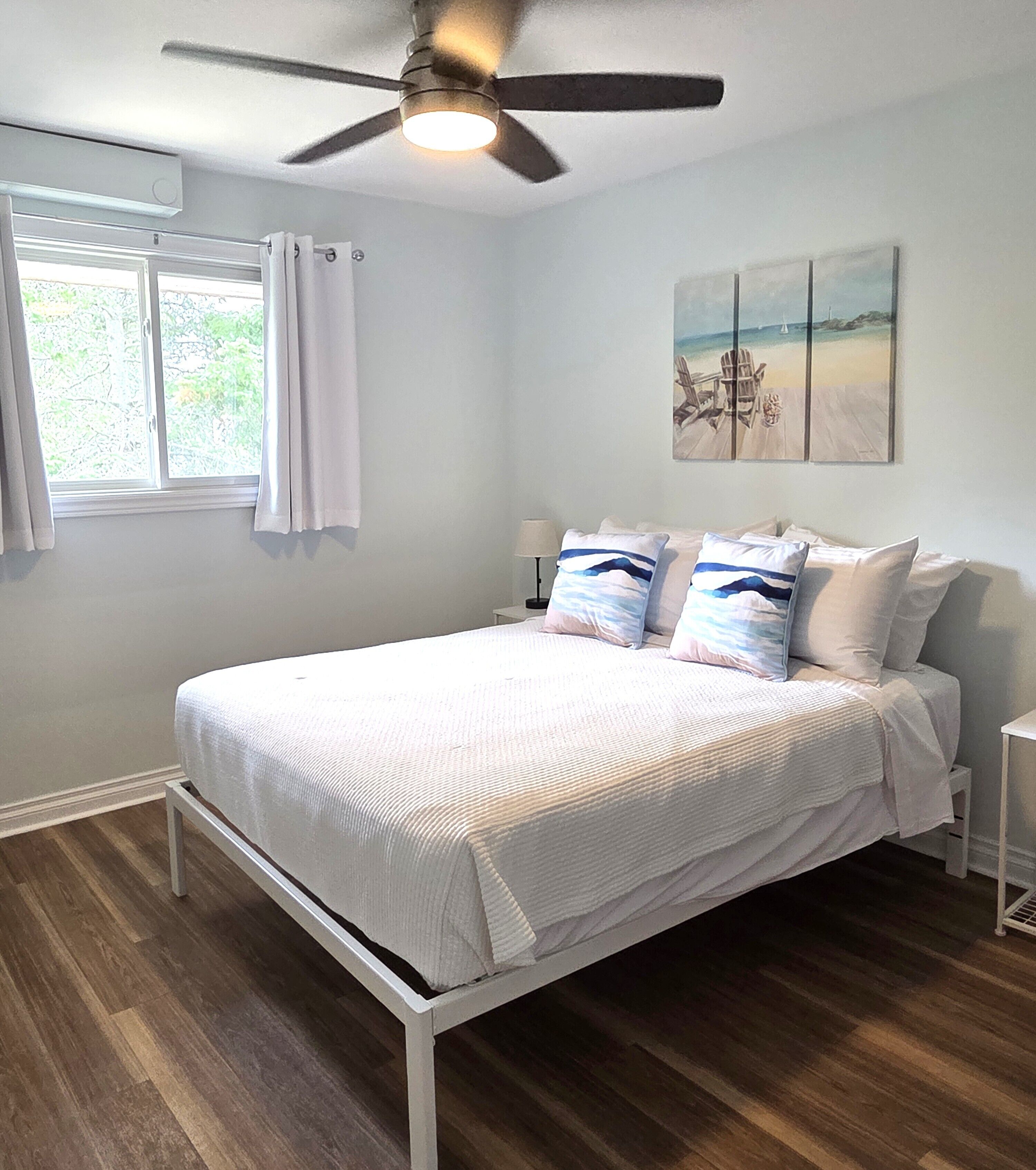 A bedroom with a bed, a ceiling fan, a window with curtains, a bedside table with a lamp, and a painting on the wall.