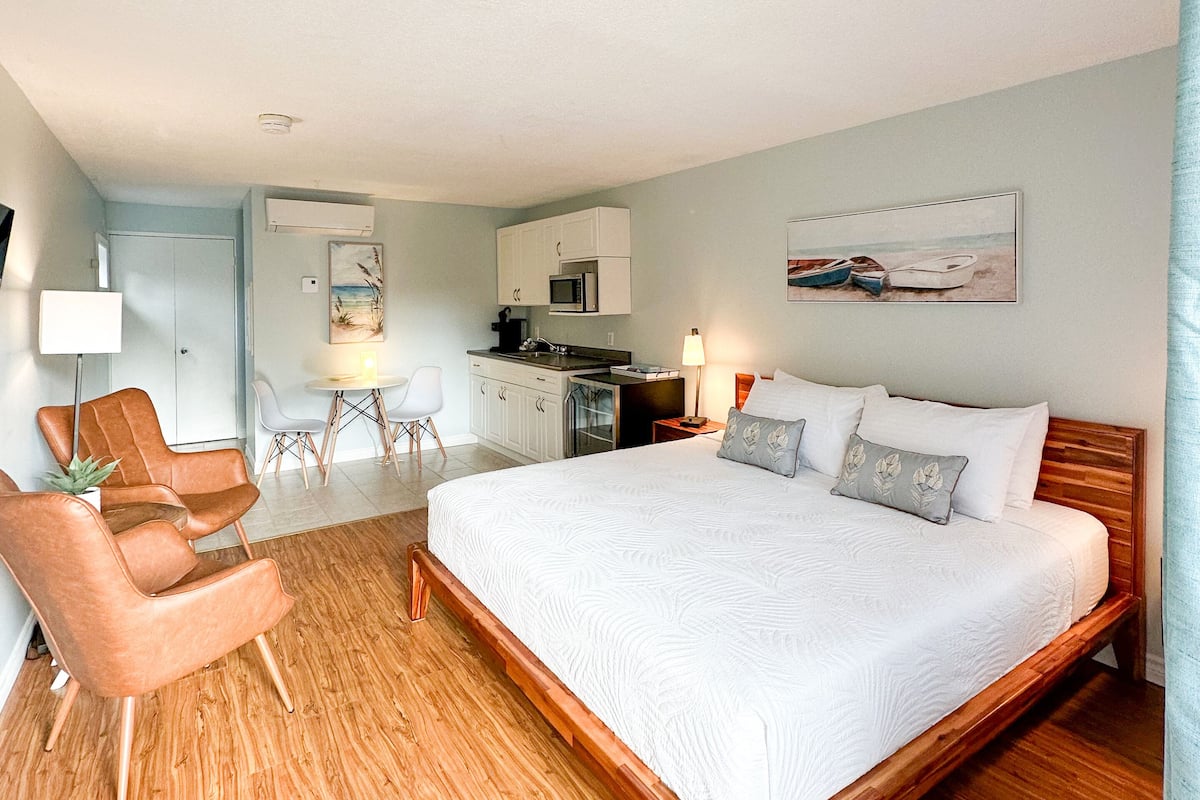 A hotel room with a large bed, wooden flooring, a kitchenette, a dining area, and a seating area with chairs.