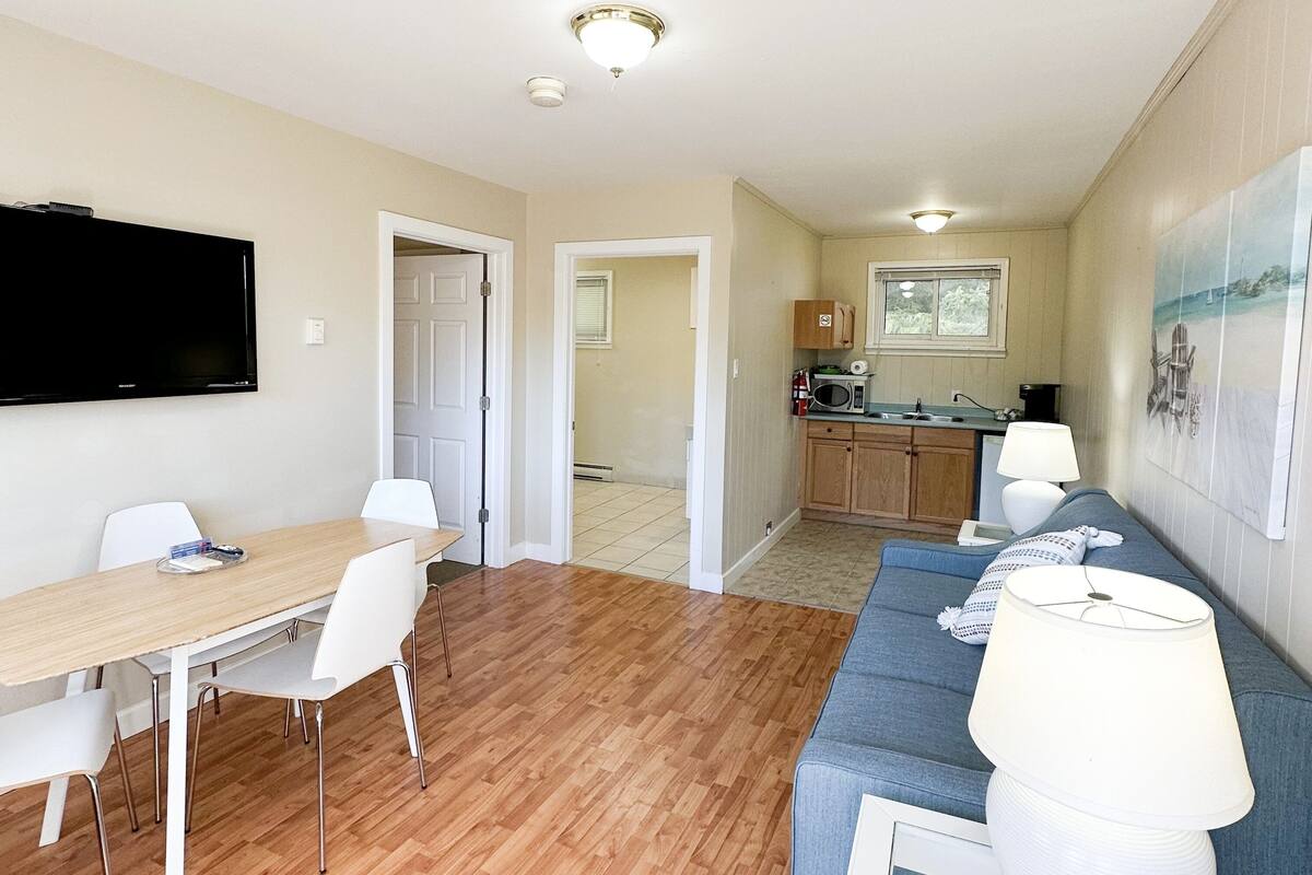 A modern living room with a wooden floor, a dining area with a table and chairs, a kitchen with a microwave, and a large TV mounted on the wall.