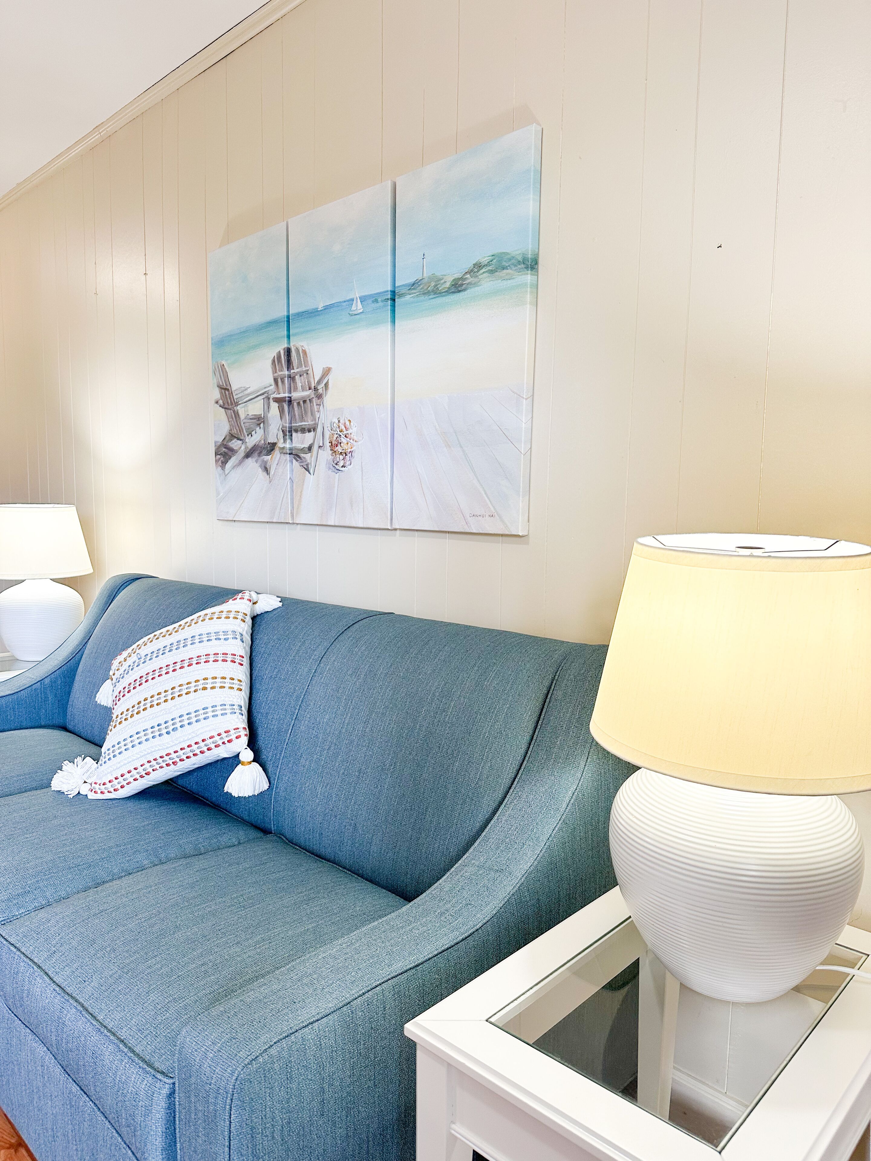 A living room with a blue sofa, a white lamp, and a painting of a beach scene.
