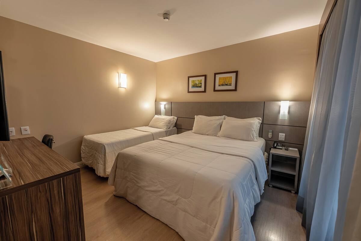 A hotel room with two single beds, a wooden desk, a nightstand with a lamp, and a small shelf.