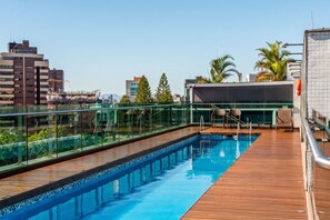 Piscina no terraço com guarda-corpo de vidro, deck de madeira e vista para a paisagem urbana.