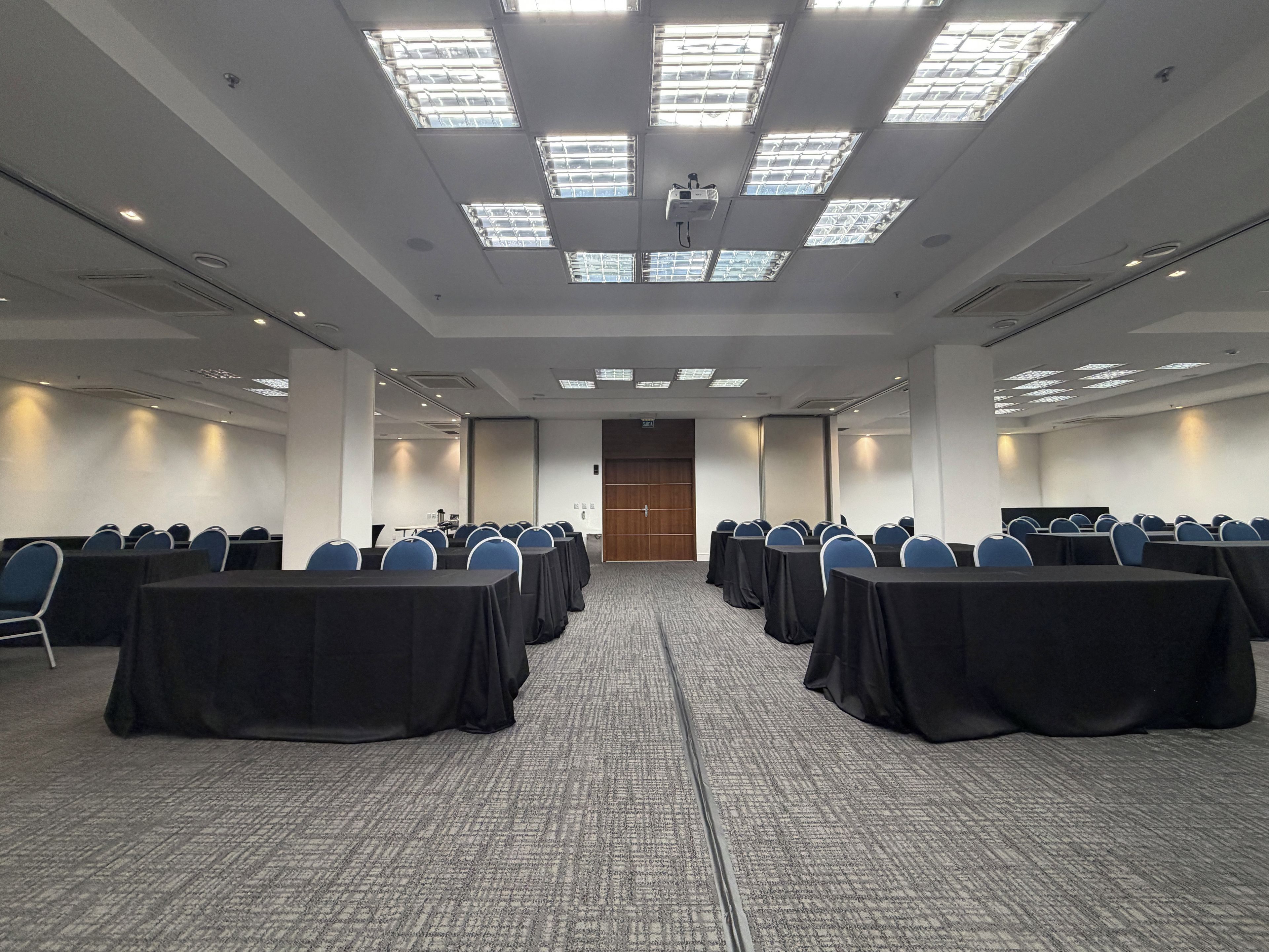 Un salón de conferencias con filas de mesas y sillas, un proyector en el techo y suelo alfombrado.