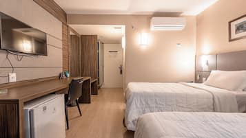 Habitación de hotel con una televisión montada en la pared, un escritorio con refrigerador, dos camas con ropa blanca y un equipo de aire acondicionado.