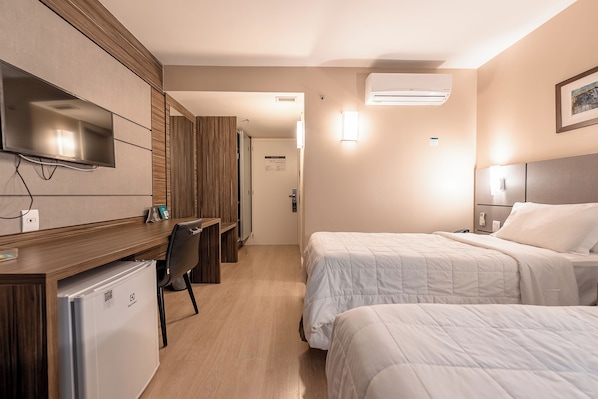 A hotel room with a TV mounted on the wall, a desk with a refrigerator, two beds with white bedding, and an air conditioning unit.