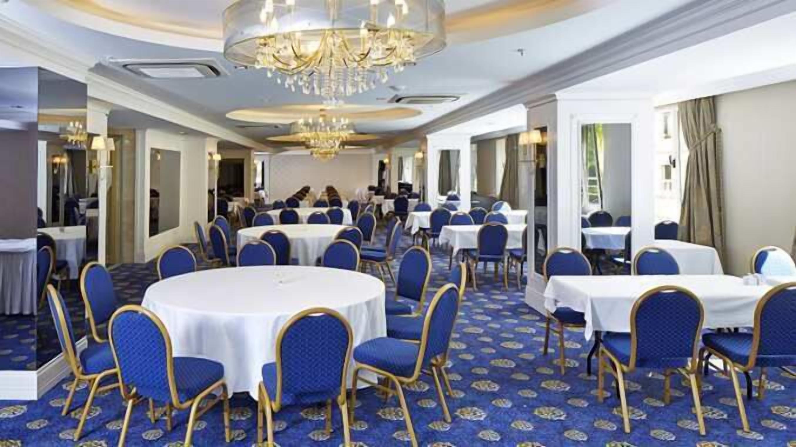 A spacious banquet hall with round tables and blue chairs, featuring a chandelier and patterned carpet.