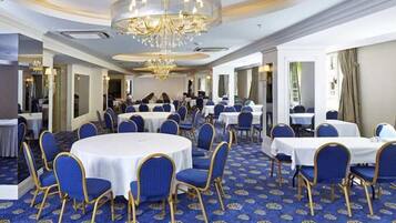 A spacious banquet hall with round tables and blue chairs, featuring a chandelier and patterned carpet.