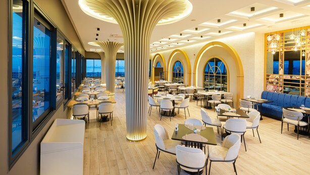 Un restaurante moderno con techos altos, amplios ventanales y una columna central. Mesas y sillas dispuestas para comer.