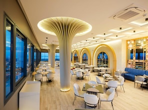 Un restaurante moderno con techos altos, amplios ventanales y una columna central. Mesas y sillas dispuestas para comer.