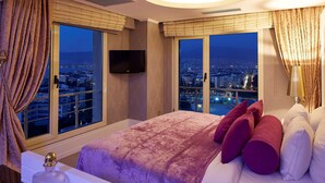 Una habitación de hotel moderna con una cama grande, dos ventanas con vistas a la ciudad, un televisor de pantalla plana y una lámpara en el techo.