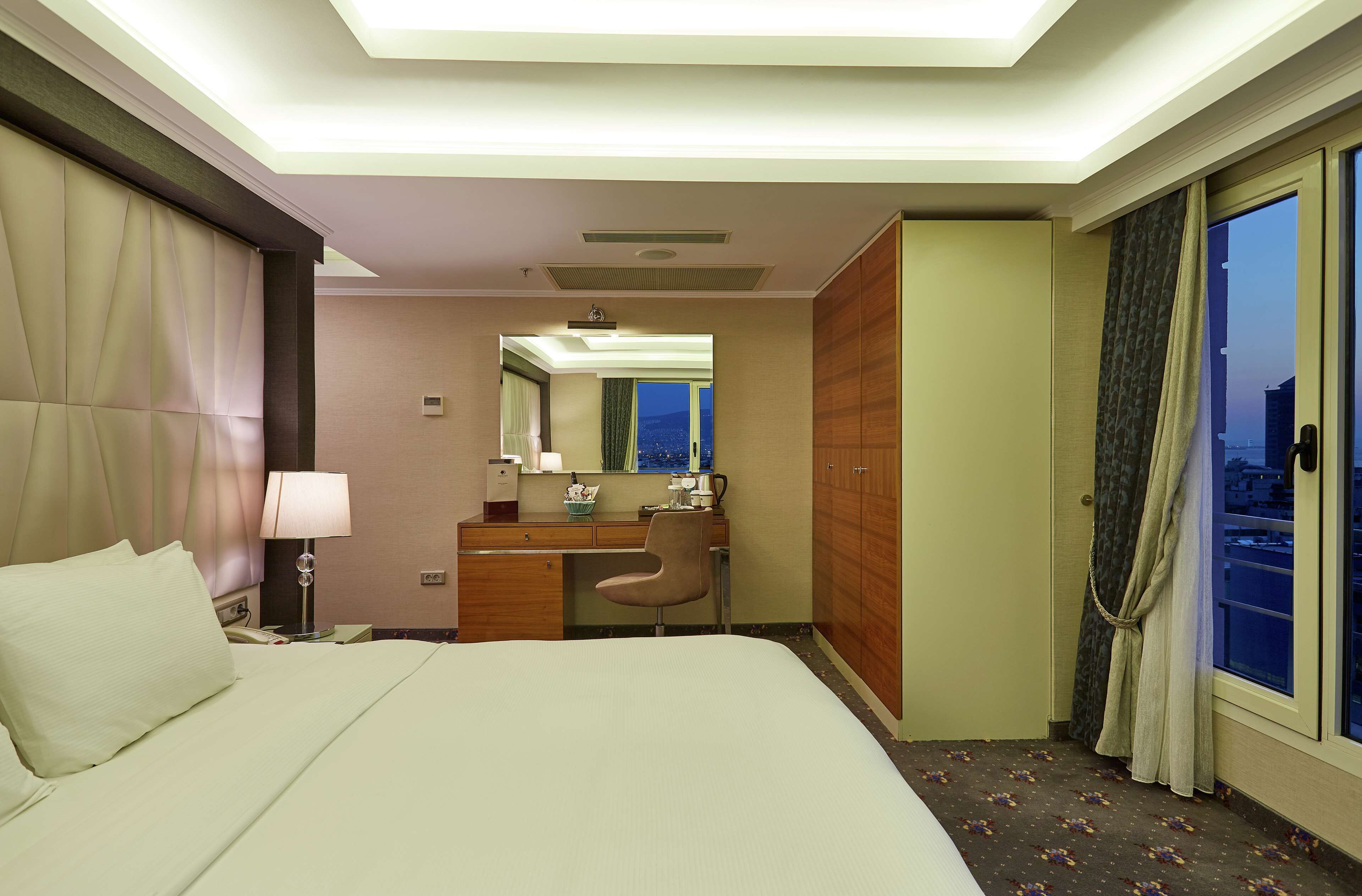 A hotel room with a bed, desk, chair, and a view of the city through the window.