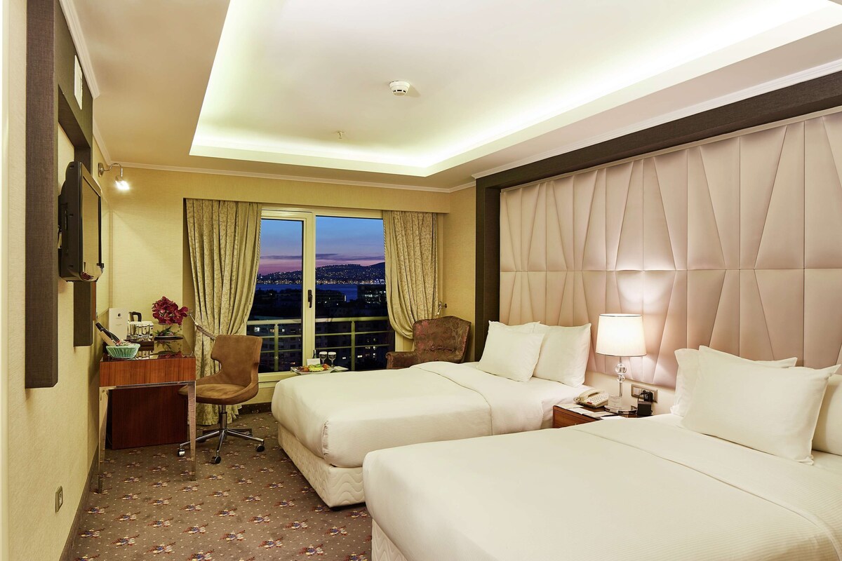 Ein Hotelzimmer mit zwei Betten, einem Schreibtisch mit Stuhl, einem Fenster mit Vorhängen und Blick auf die Stadt im Abenddämmerung.