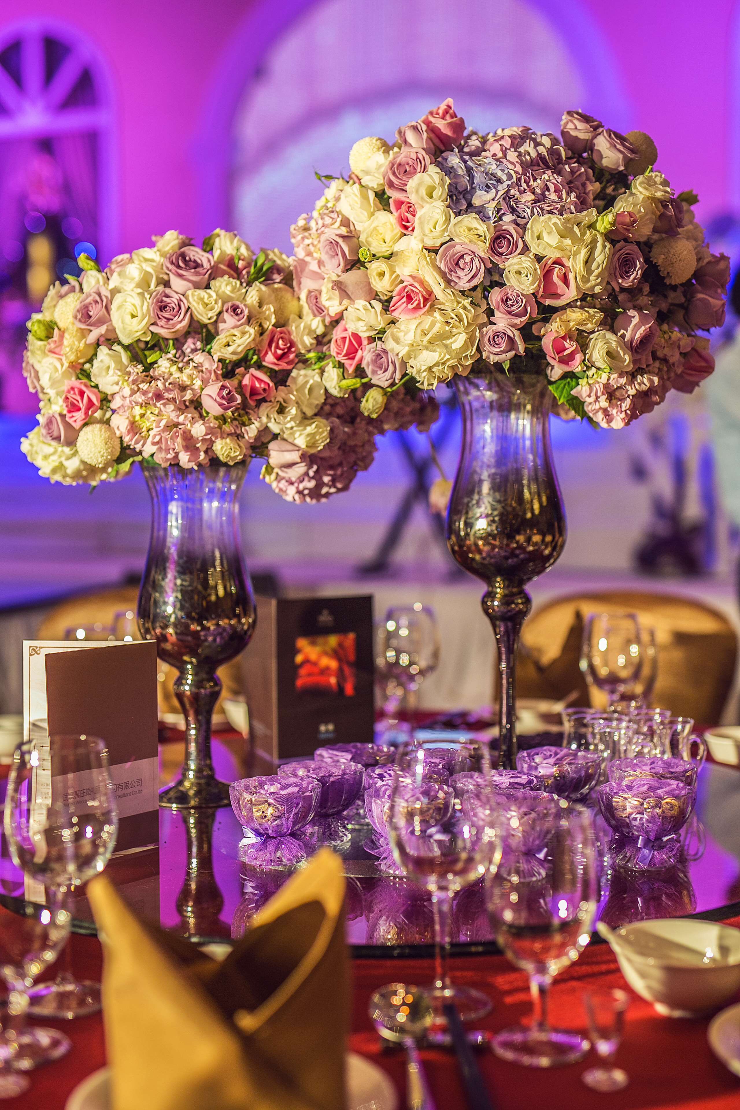 A table set for dining with two large floral arrangements in tall vases, surrounded by empty wine glasses and a menu card.