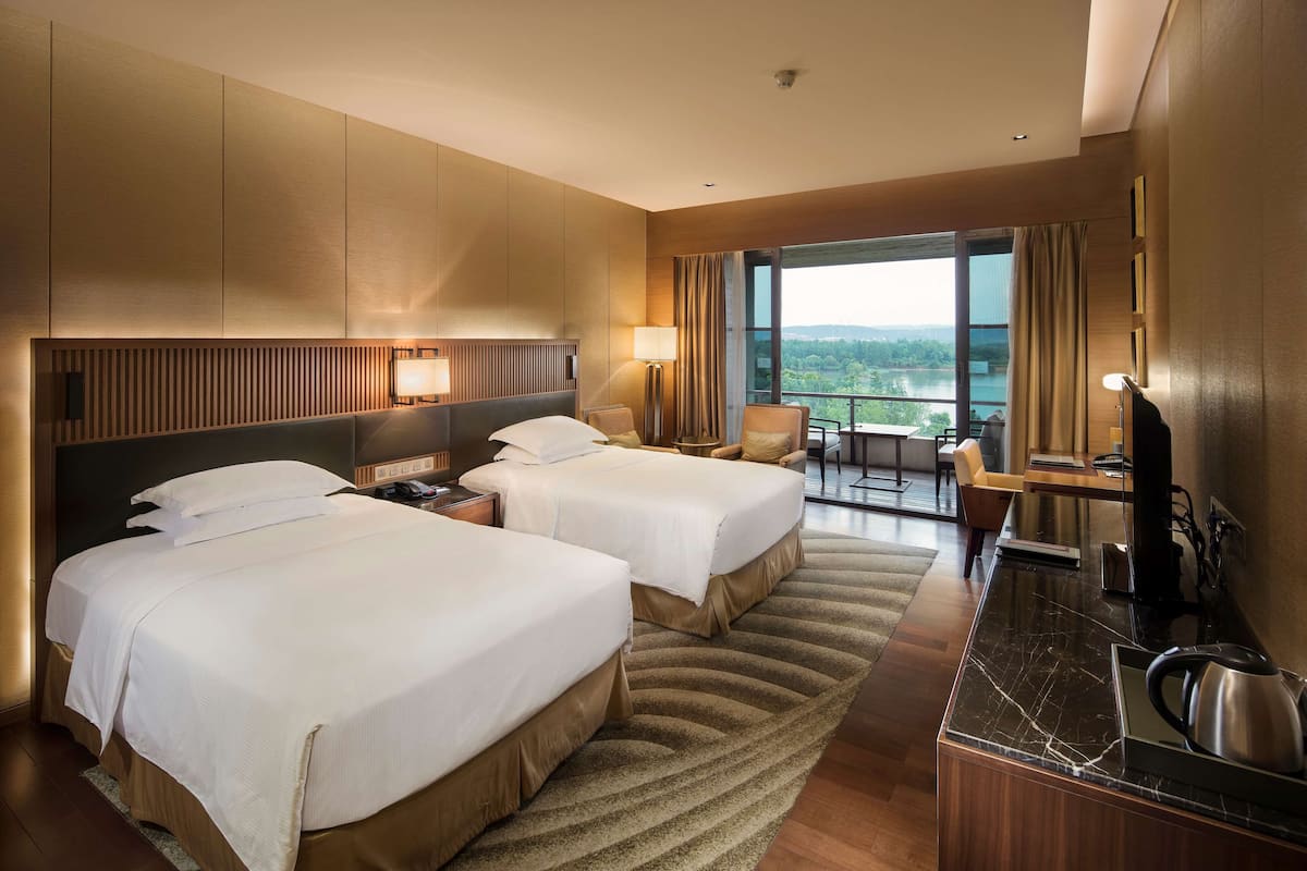 A hotel room with two beds, a desk with a chair, a large window with a view, and a balcony.