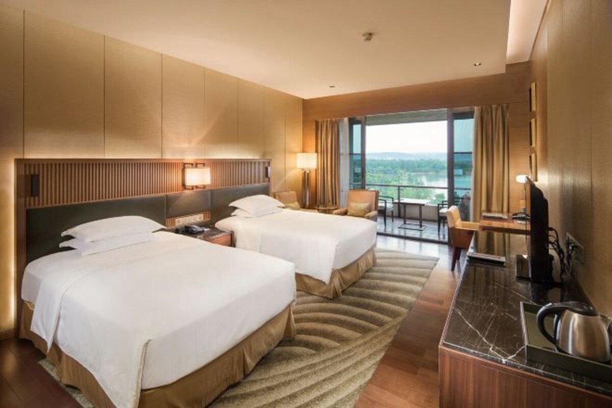 A hotel room with two beds, a desk, a chair, and a large window offering a view.