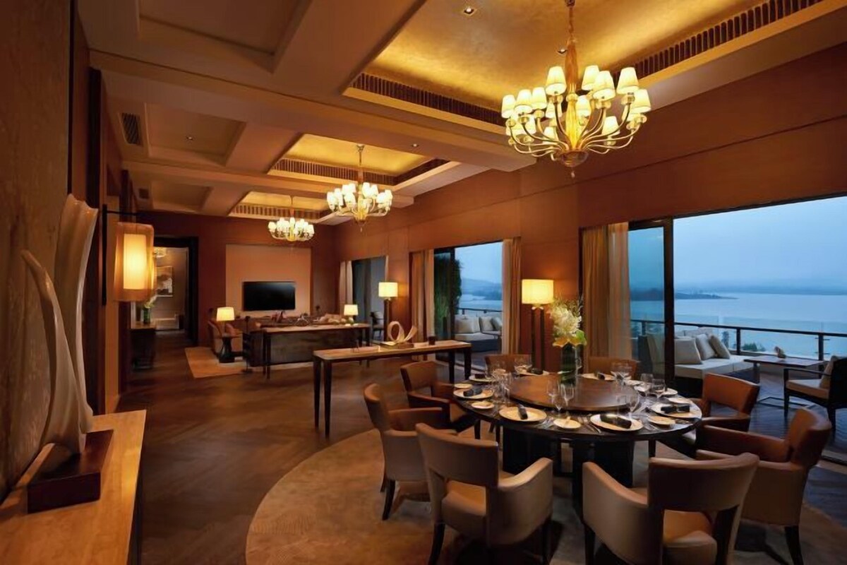 A spacious dining area with a large round table, multiple chairs, and a chandelier. There is a television and seating area in the background.