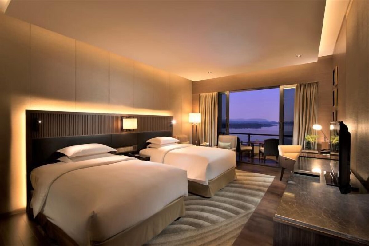 A hotel room with two beds, a desk, a chair, and a large window with a view.