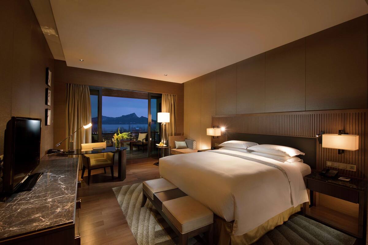 A hotel room with a large bed, a desk with a chair, a TV, and a view of the mountains.