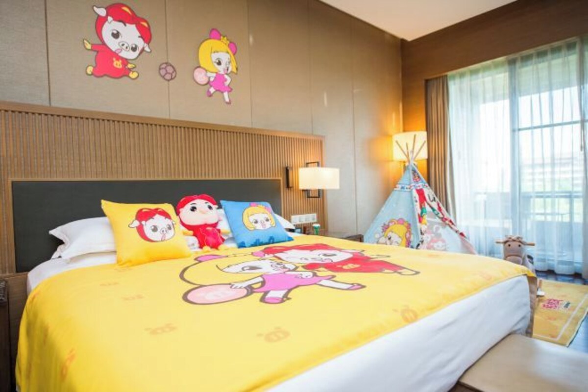 A hotel room with a large bed featuring cartoon character bedding, a wooden headboard, a nightstand with a lamp, and a window with curtains.