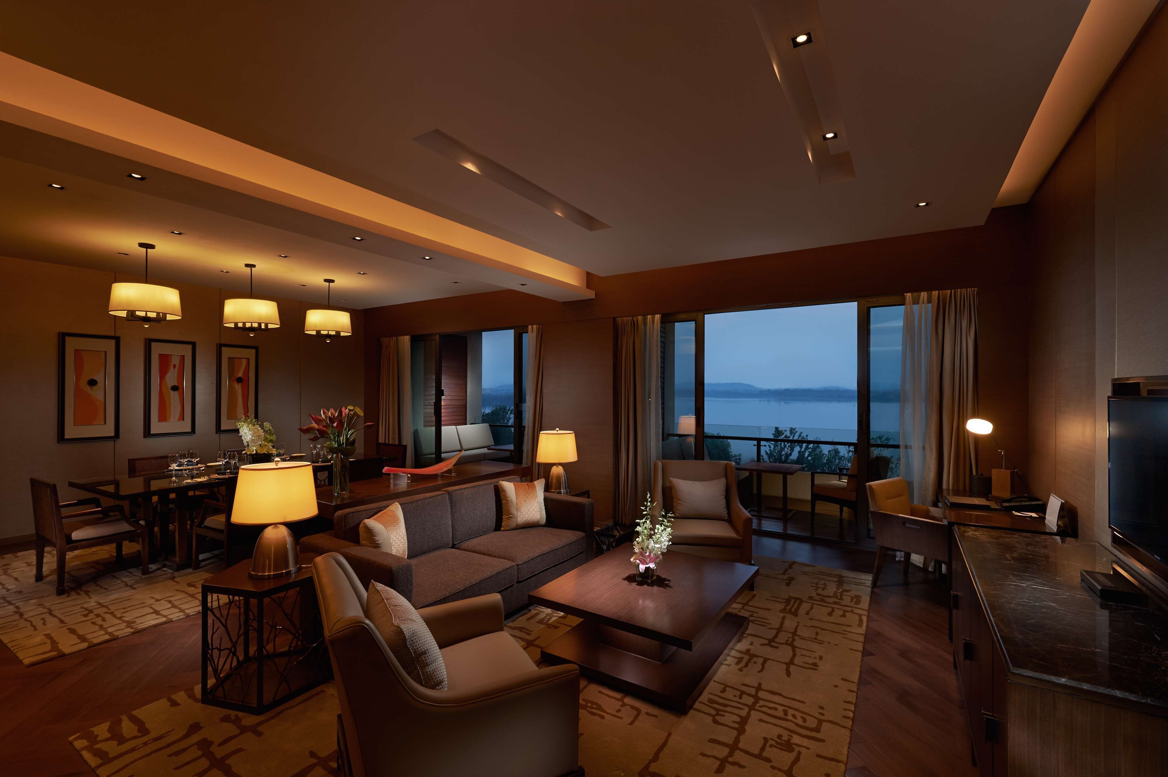 A modern hotel room with a sofa, armchairs, a dining table, and a large TV.