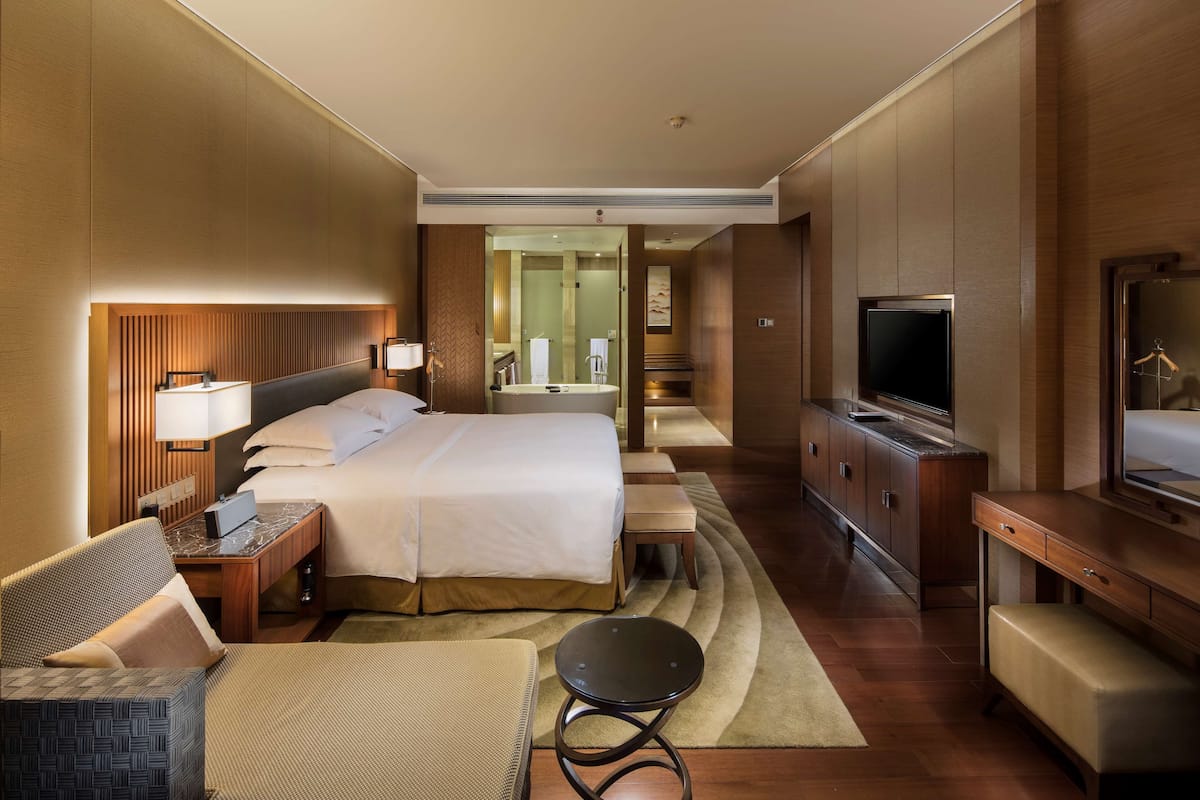 A hotel room with a bed, bedside table, lamp, TV, and a seating area with a chair and ottoman.