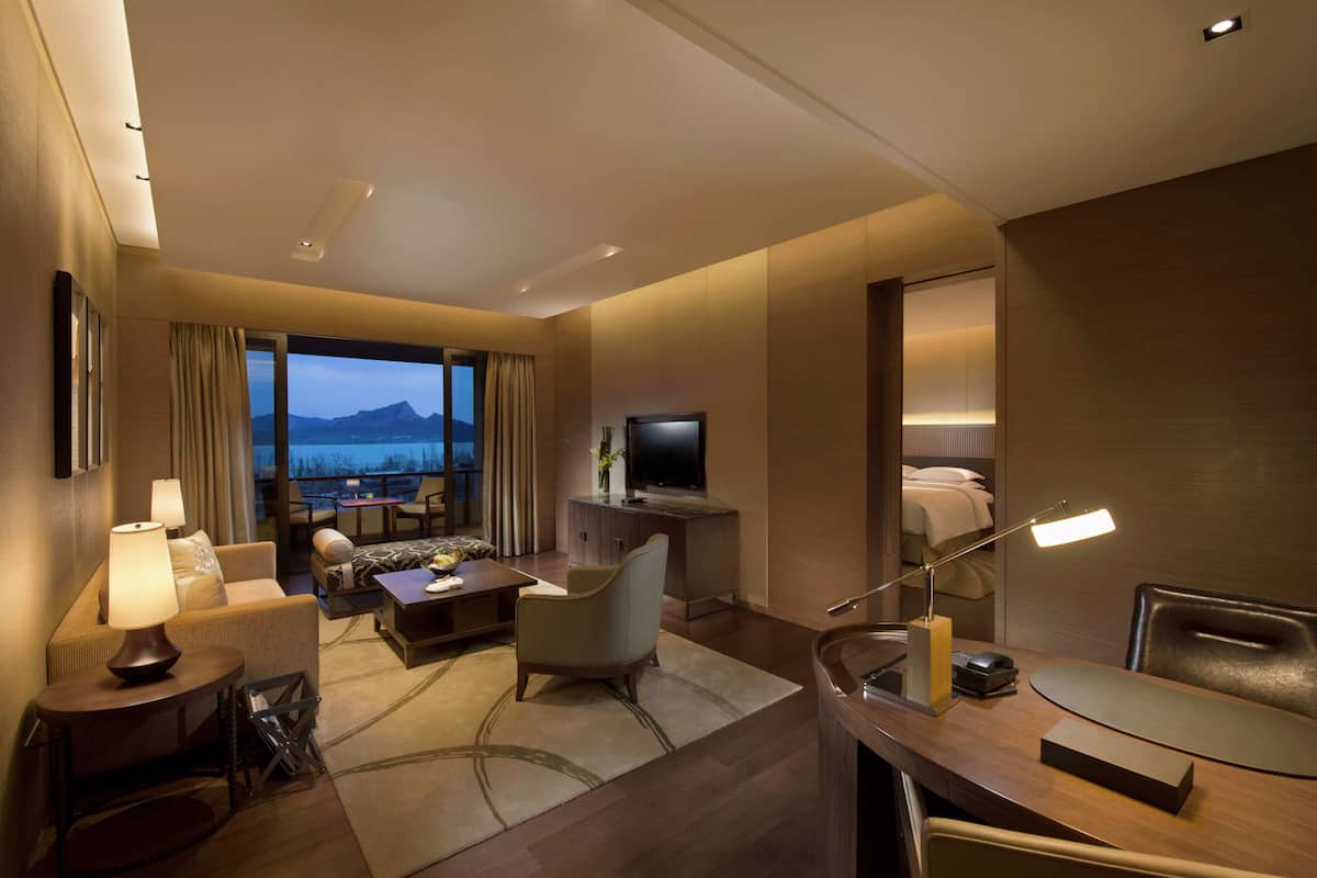 A modern hotel room with a large window, a flat-screen TV, a desk with a lamp, a sofa, and a chair.