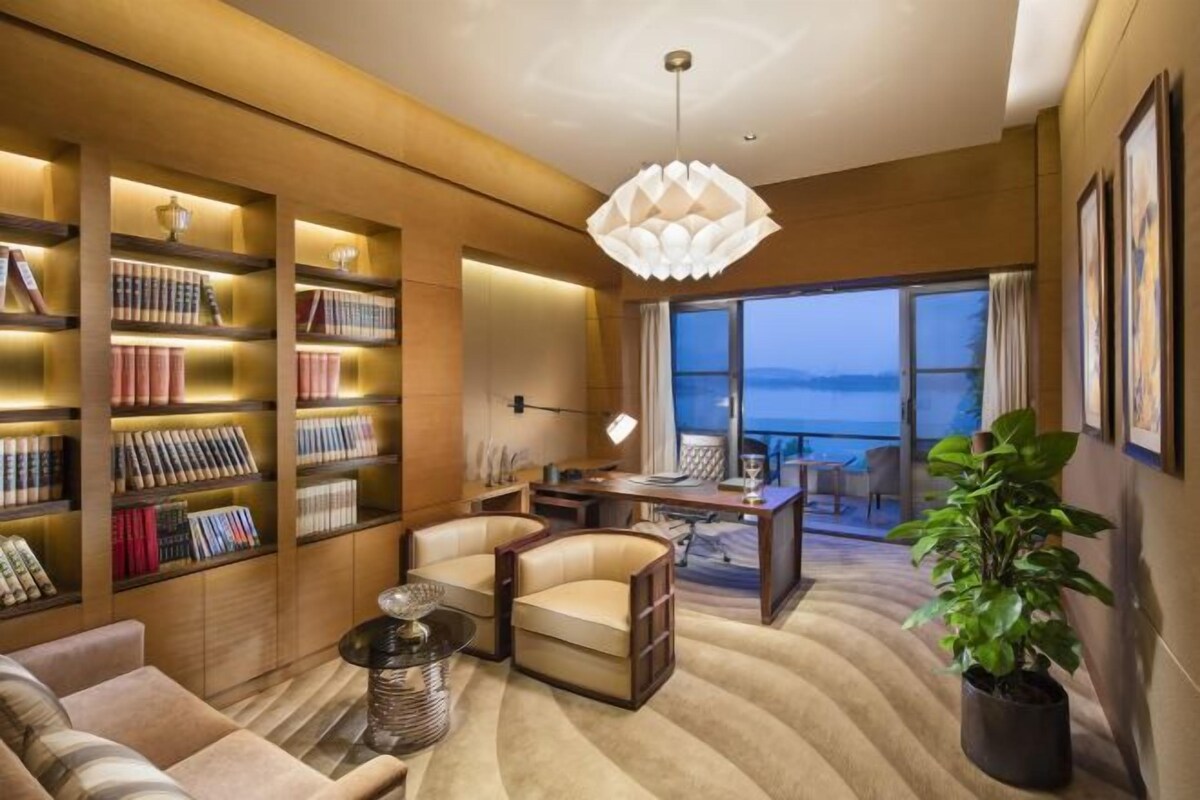 A modern hotel room with a large bookshelf, comfortable seating, a dining area, and a view of the sea.