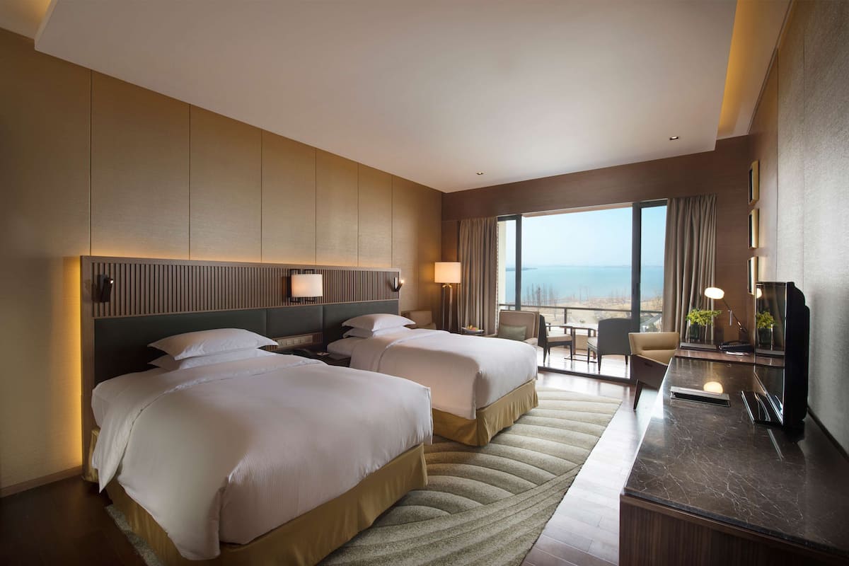 A hotel room with two beds, a desk, a chair, a TV, and a large window with a view of the sea.