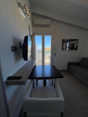 A modern living room with a dining table, a sofa, a TV mounted on the wall, and a large window offering a view of the outdoors.