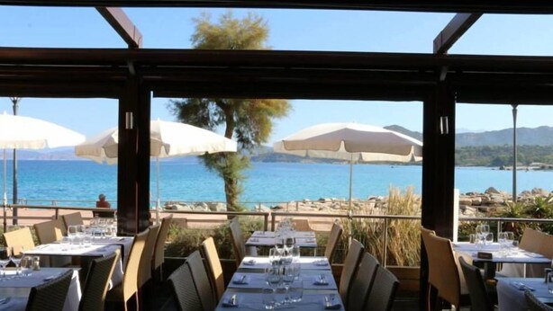 An outdoor dining area with tables set for two, overlooking a body of water and mountains. - Hôtel L'Escale Coté Port (L'Île-Rousse)
