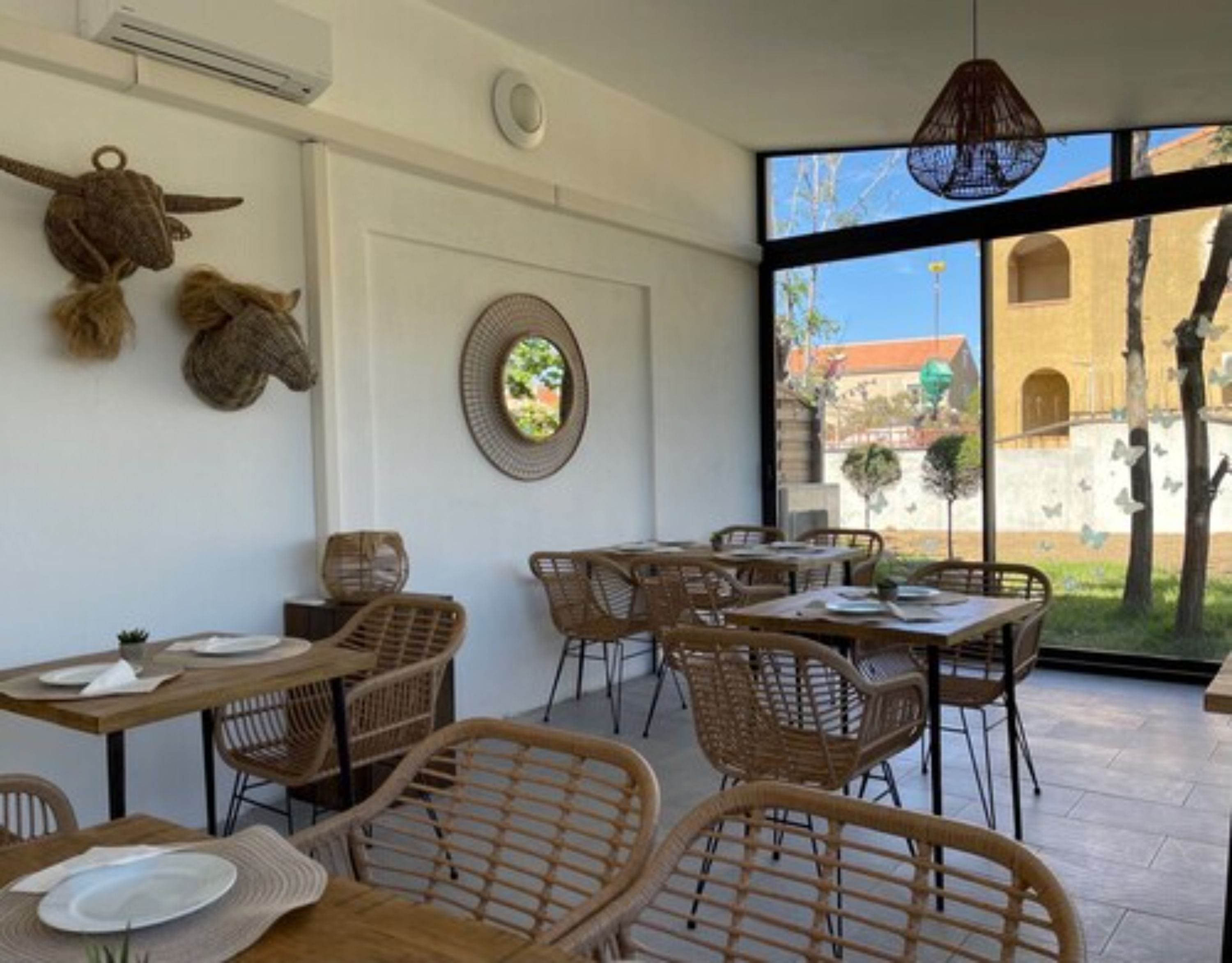 Un restaurante con mobiliario de mimbre, un espejo redondo y vistas a un edificio a través de amplios ventanales.
