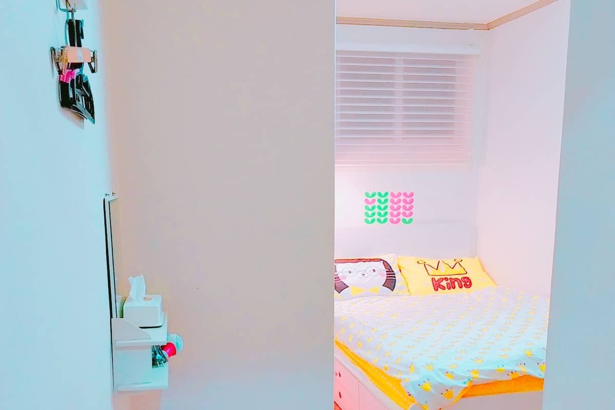 A bedroom with a bed, a small stool, a white cabinet, and a window with blinds.
