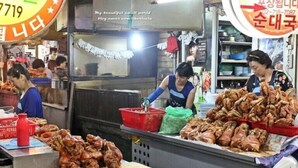 Un étal de marché où diverses viandes sont exposées sur des tables, une enseigne avec du texte coréen, et une personne qui prépare de la nourriture.