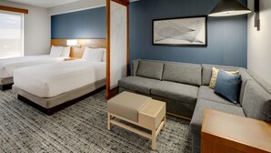 Ein Hotelzimmer mit zwei Betten, einem Sofa, einem kleinen Tisch, einer Lampe und einem gerahmten abstrakten Gemälde an der Wand.