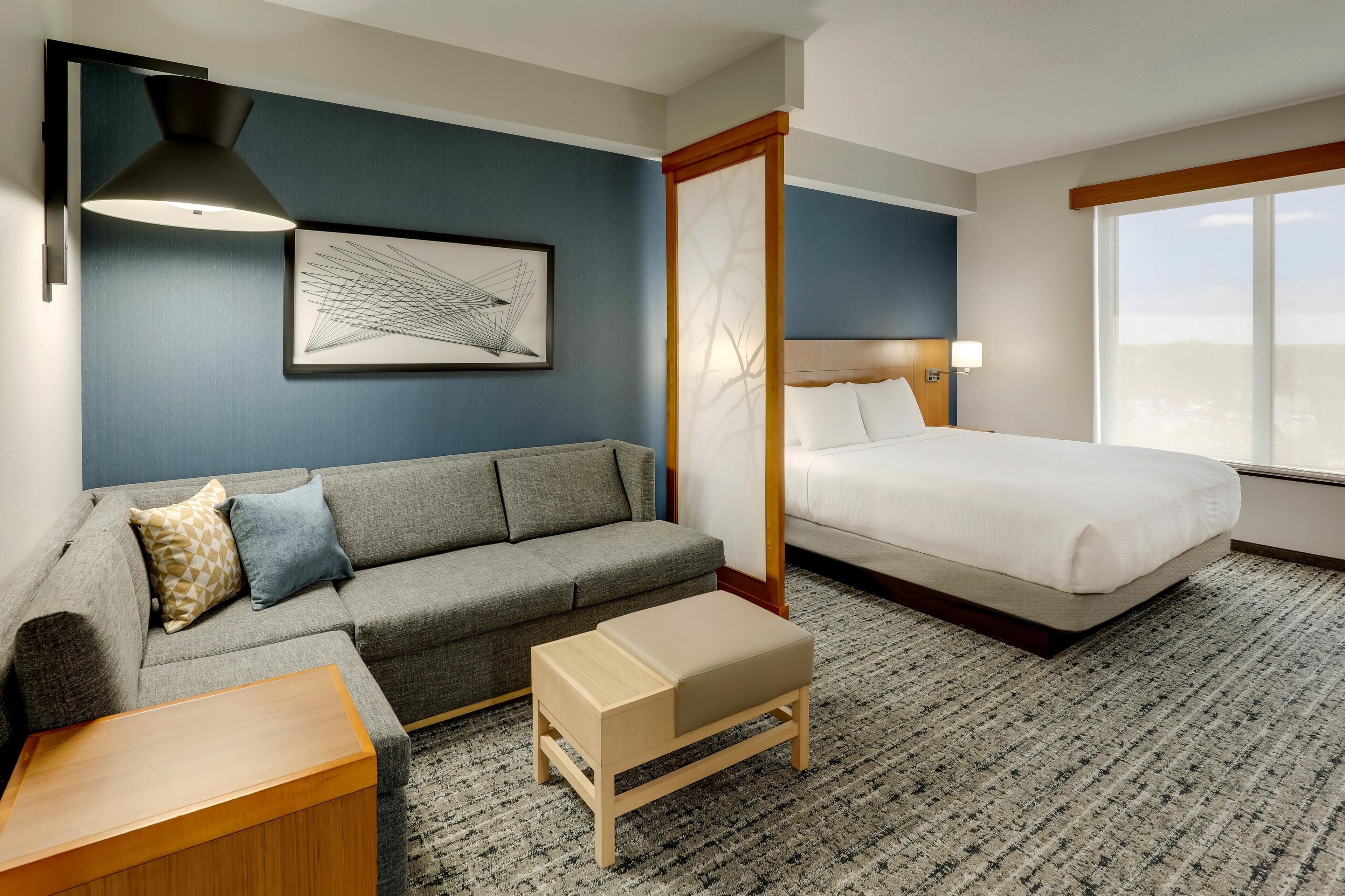 Une chambre d’hôtel avec un lit, un canapé, une petite table en bois, une lampe sur pied et une grande fenêtre.