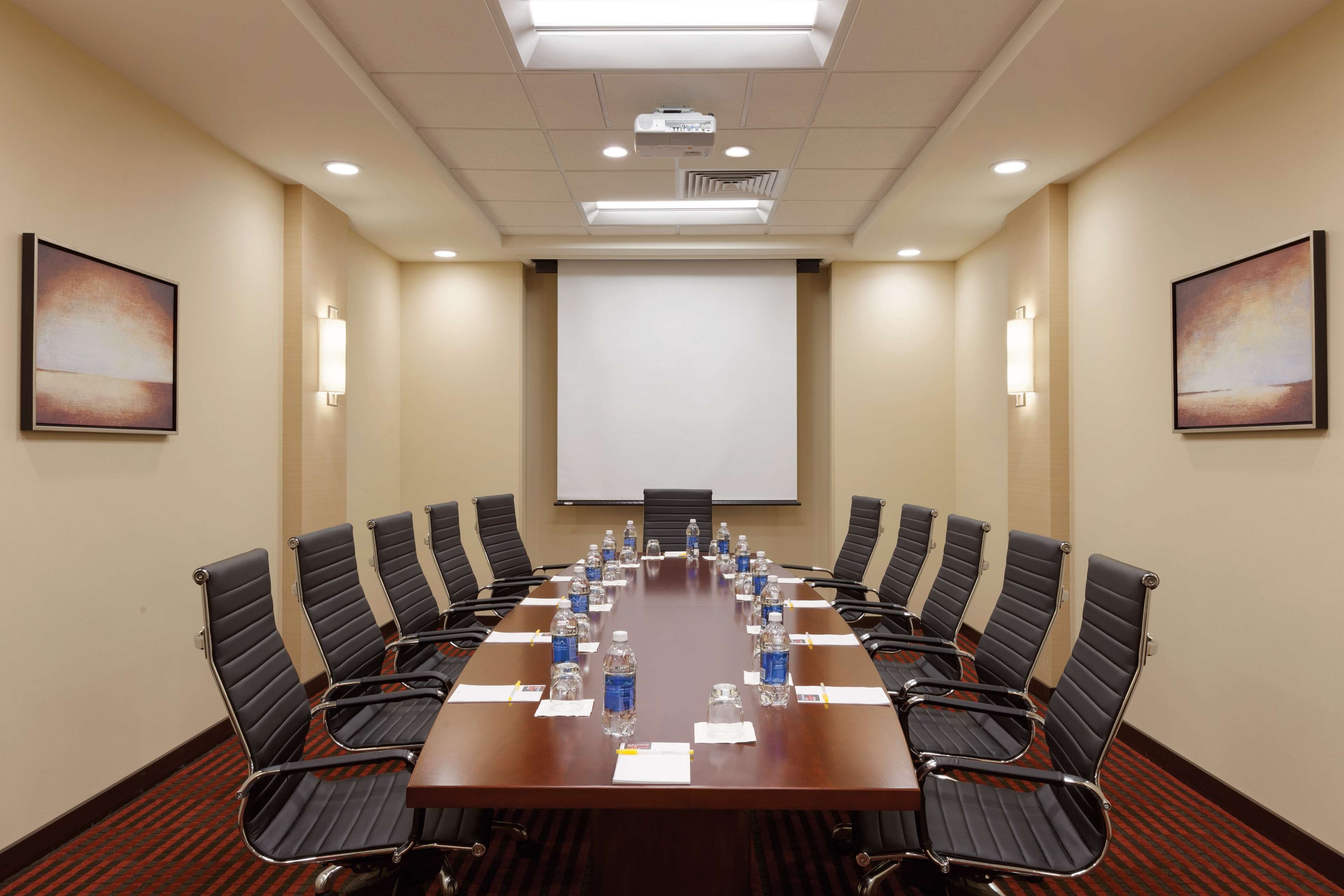 Un salón de conferencias con una larga mesa, sillas negras, botellas de agua y una pantalla para proyector.