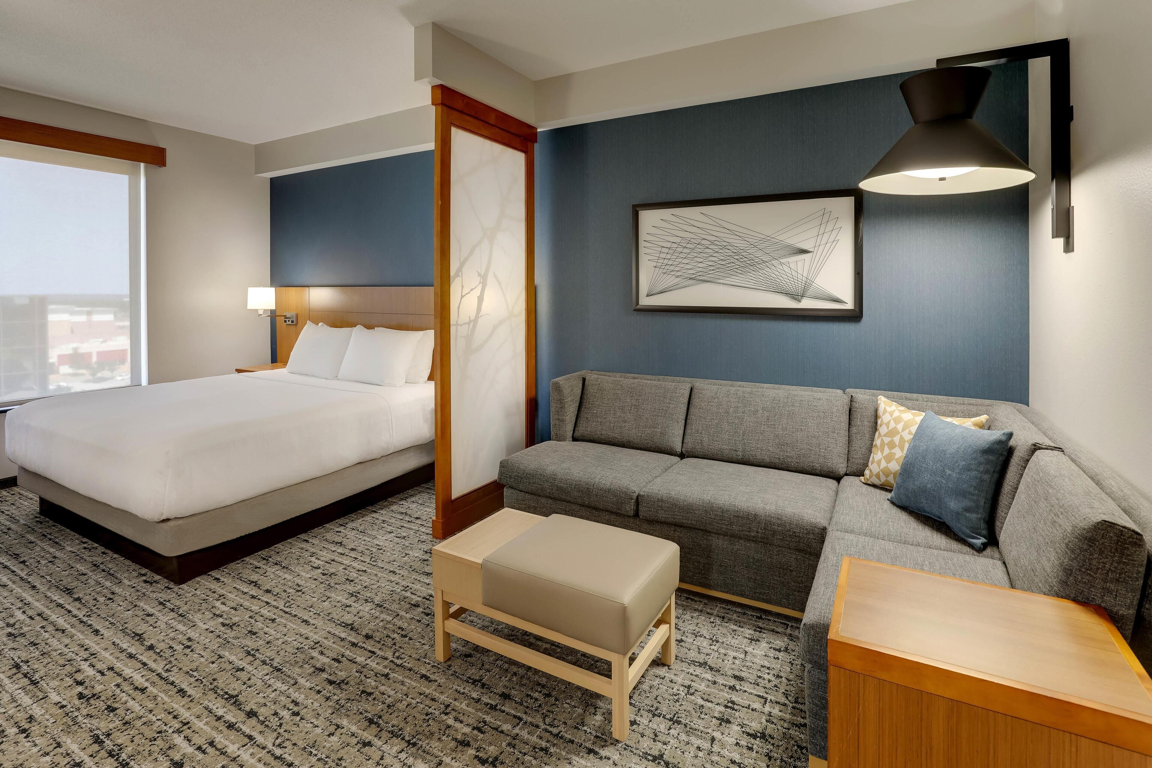 Een hotelkamer met een bed, een bank, een voetenbankje, een bijzettafeltje, een muurlamp en een lijst met abstracte kunst.