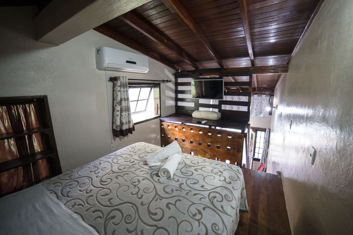 Un dormitorio con una colcha estampada, un cabecero de madera, una ventana con cortinas y un televisor sobre una cómoda de madera.