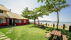 A red building with a tiled roof, a grassy lawn, picnic tables, and a view of the sea with boats. - Pousada dos Ingleses (Bombinhas)