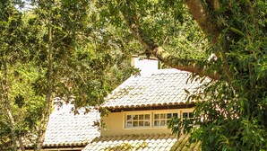 A two-story building with a red facade and a tiled roof, surrounded by lush greenery and a wooden bridge. - Pousada dos Ingleses (Bombinhas)