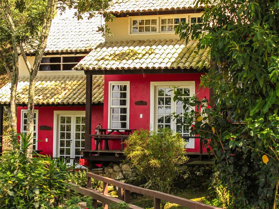 A two-story building with a red facade and a tiled roof, surrounded by lush greenery and a wooden bridge.