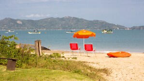 A beach with red chairs and an orange umbrella, boats in the water, and mountains in the background. - Pousada dos Ingleses (Bombinhas)