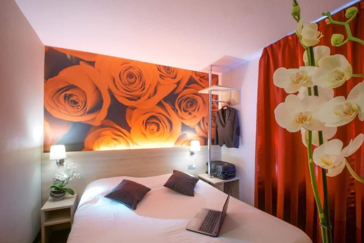 A hotel room with a bed, a laptop, a nightstand with a lamp, a wall with a floral mural, and a red curtain.