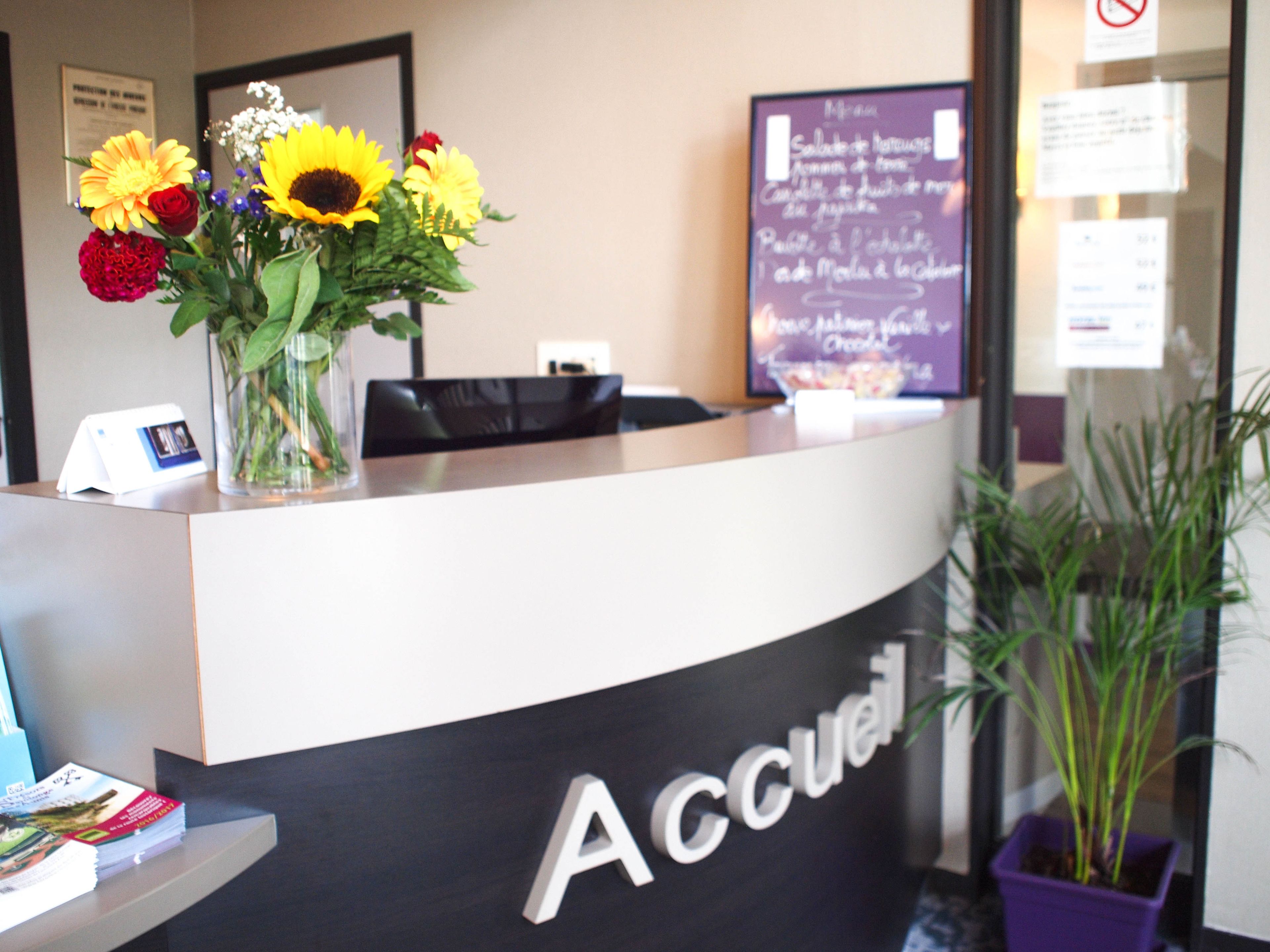 A reception desk with a vase of flowers, a sign reading "Accueil", and a menu board in the background.