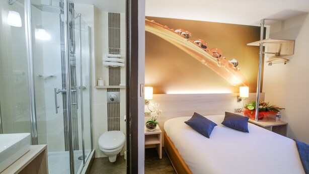 A hotel room with a glass-enclosed shower, a toilet, a bed with two pillows, a nightstand with a lamp, and a wall-mounted shelf with a plant. - Hôtel Inn Design & Restaurant Rochefort (Rochefort)
