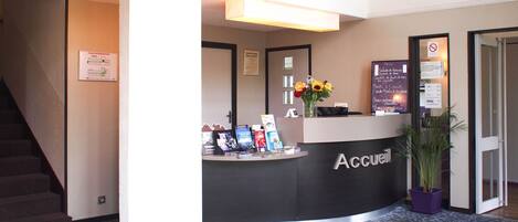 A reception desk with a sign reading "Accueil" and a flower vase. There are brochures and a menu on the desk.