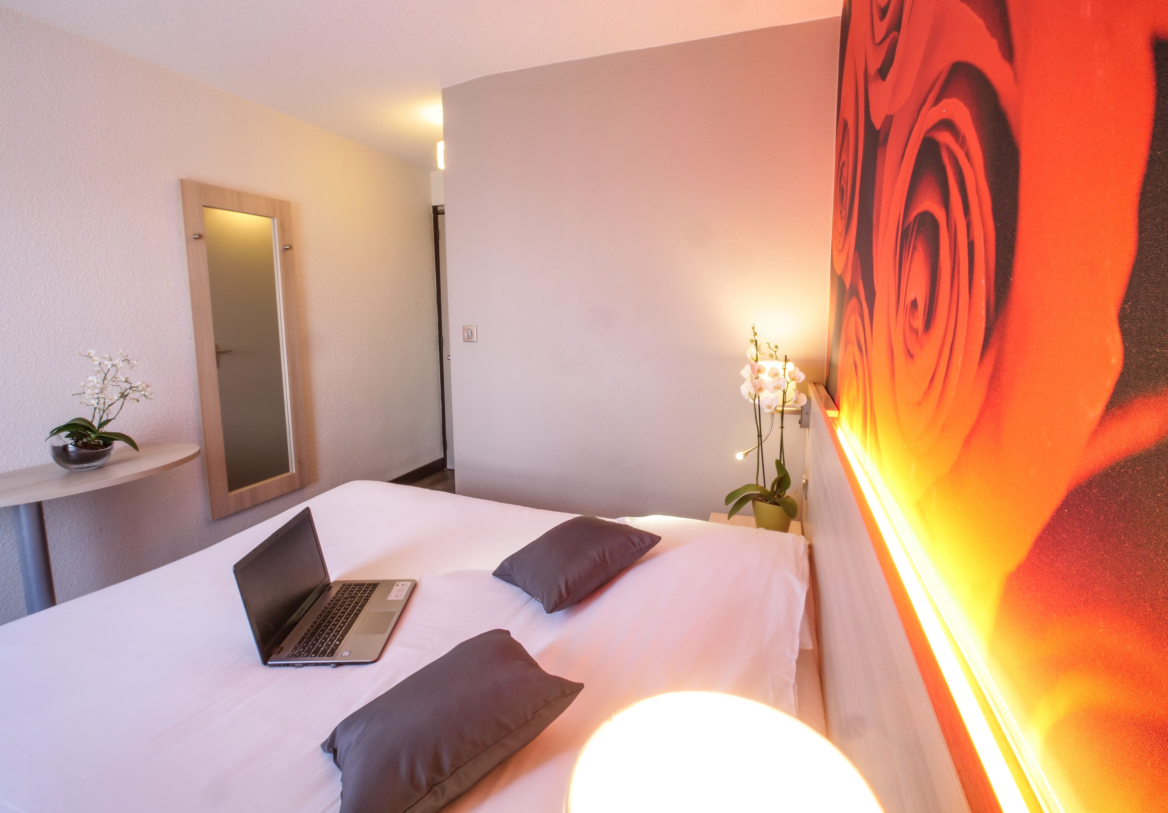 A hotel room with a bed, a laptop, a mirror, and a large floral painting on the wall.