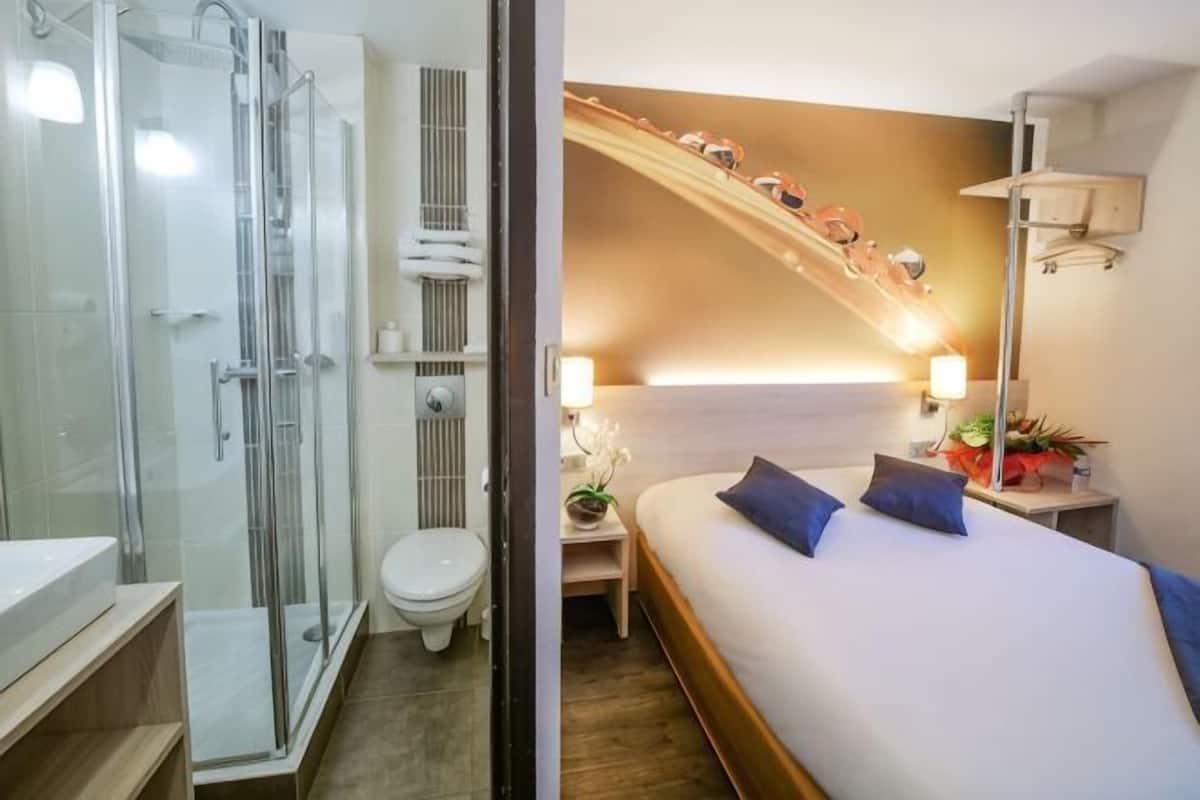 A hotel room with a glass shower enclosure, a bed with blue pillows, a bedside table with a lamp, and a wall-mounted shelf with decorative items.