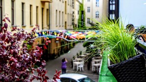Une rue étroite bordée de bâtiments des deux côtés, un pont piétonnier illuminé de lumières colorées et un pot de fleurs au premier plan.