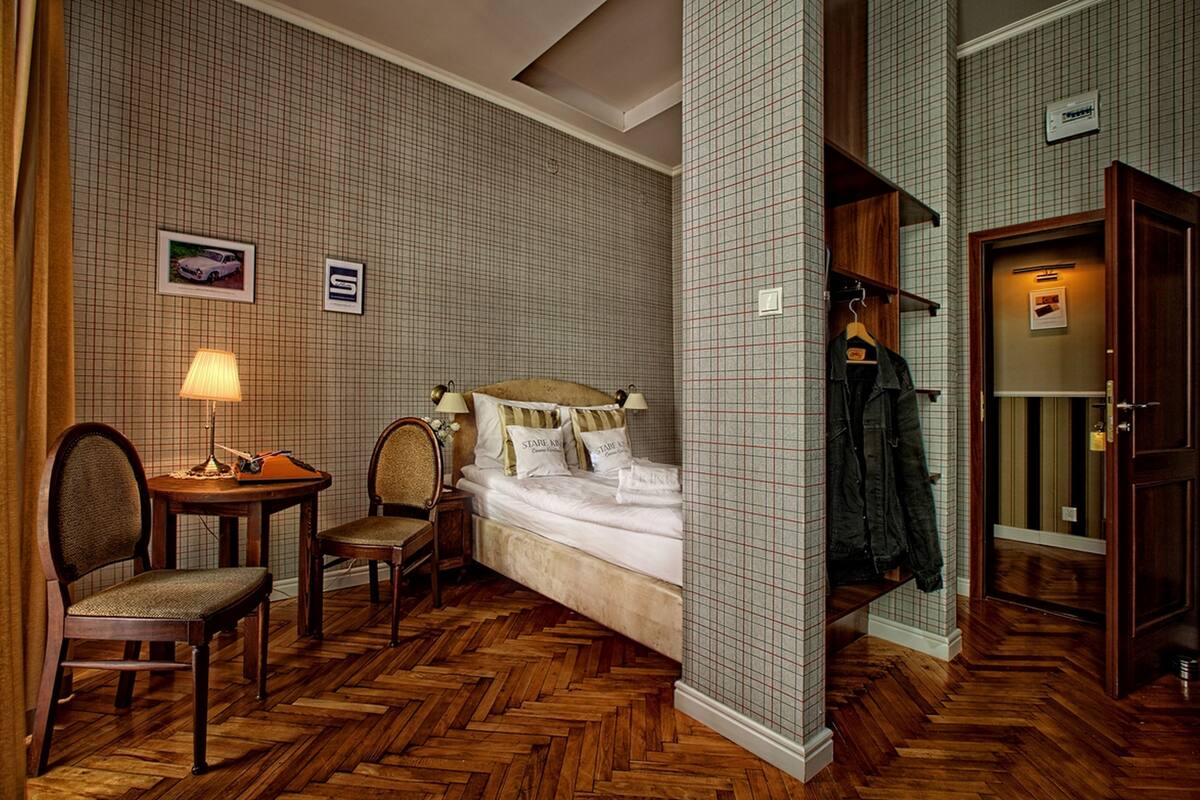 Ein Hotelzimmer mit einem Bett, zwei Sesseln, einem Tisch mit Lampe, einem Kleiderständer und einem Holzboden mit Fischgrätmuster.
